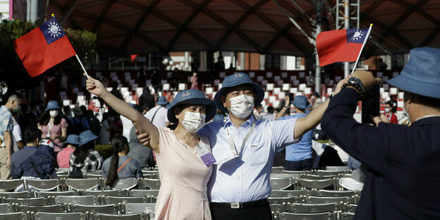 Ein Paar mit taiwanesischen Fähnchen bei einer Veranstaltung. Sie tragen Maske. Im Hintergrund andere Menschen, die mit dem Rücken zum Paar auf Klappstühlen sitzen.