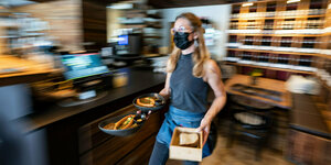 Eine Kellnerin mit Maske trägt in einem Restaurant Essensteller
