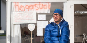 Im Vordergrund ist ein Mann mit blauer Mütze und blauer Jacke zu sehen. Im Hintergrund stehen an einer gläsernen Wand die Worte "Hungerstreik" geschrieben.