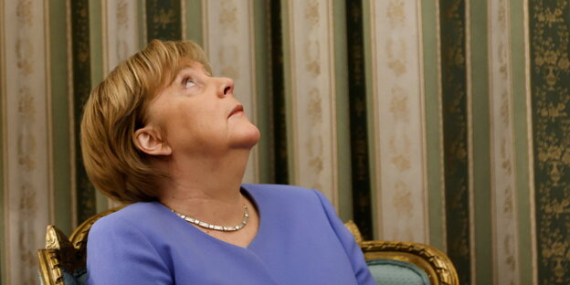 Merkel sitzt auf einem goldenen Stuhl und schaut nach oben, hinter ihr eine grün-goldene Tapete