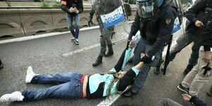 Ein Mann liegt auf dem Asphalt einer Straße und wird von einem uniformierten Polizisten weggeschleift, mehrere Menschen stehen drumherum