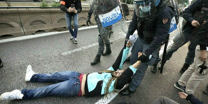 Ein Mann liegt auf dem Asphalt einer Straße und wird von einem uniformierten Polizisten weggeschleift, mehrere Menschen stehen drumherum