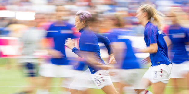 Megan Rapinoe rennt zwischen MItspielerinnen auf dem Fußballfeld. Das Bild ist unscharf aufgrund der Bewegung