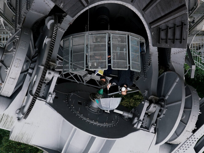 Umgekehrtes Bild des Sitzplatz am Fernrohr in der Archenhold Sternwarte, ein Mann mit verdunkelter Brille liest ein Buch, hinter ihm hält ein zweiter Mann einen Regenschirm über ihn