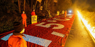 Mehrere Personen stehen vor einem Protest-Banner, dahinter brennt eine Linie aus Feuer