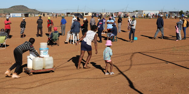 Kinder schleifen Wassercontainer über die Erde, dahinter eine Schlange von Wählern