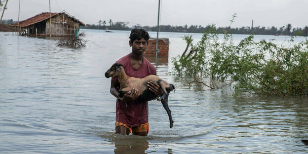 Ein Junge steht mit den Beinen im Hochwasser, er rettet eine Ziege in seinen Armen