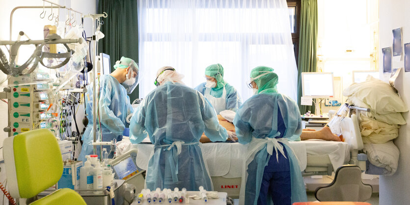 Blick in ein Patientenzimmer in einem Krankenhaus. Um ein Bett herum stehen mehrere Menschen, die Schutzkleidung anhaben