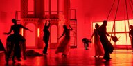 Die Bühne ist in rotes Licht getaucht, als Schatten erscheinen Männer und Frauen in historisierenden Kostümen.