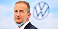 VW-Chef Diess mit unter das Kinn gezogener Atemmaske vor einem Logo des Volkswagen-Konzerns