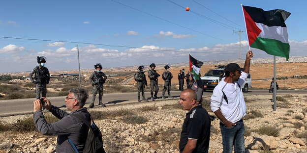 Demonstranten mit einer palästinensichen Flagge und Uniformierte in karger Landschaft.