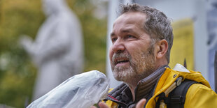 Sören Benn steht in gelber Regenjacke vor einer Kirche, im Hintergrund ein Steinengel