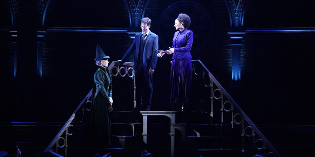 Auf einer dunklen Bühne steht eine Treppe, darauf stehen beleuchtet drei Schauspielerinnen und Schauspieler