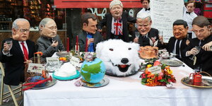 Als Politiker verkleidete Menschen sitzen an einem Tisch um einen Eisbärkopf herum.