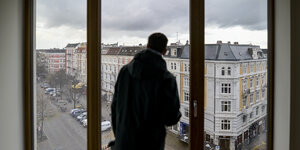 Ein Mann steht in einer Wohnung und sieht aus dem Fenster auf die gegenüberliegenden Häsuer.