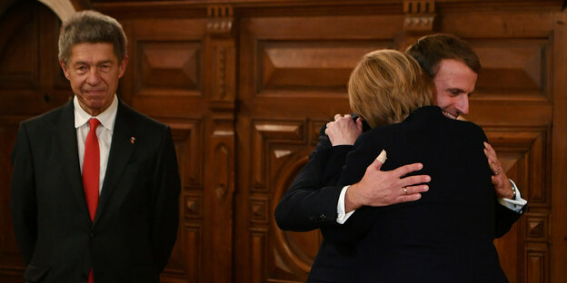 Angela Merkel umarmt Macron, Ehemann Sauer steht daneben.