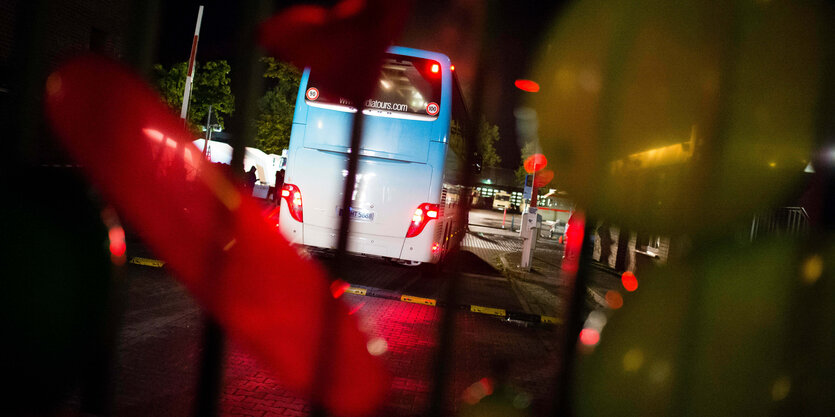 Die Rücklichter eines Reisebusses leuchten hinter einem Zaun und einem Schlagbaum.