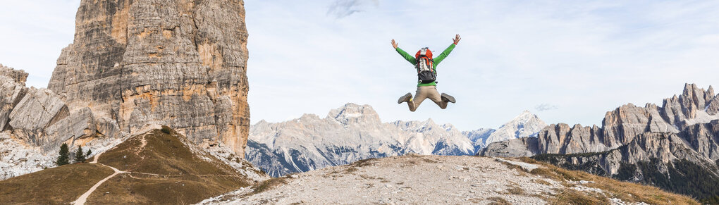 In einem Gebirge springt eine Person mit Kletterausrüstung vor blauem Himmel in die Luft