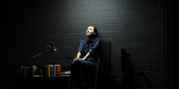 Eine Frau sitzt vor einer schwarzen Wand und schaut bedrückt in die Ferne.