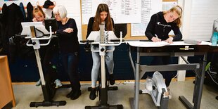 Schülerinnen treten auf Ergometern und machen dabei Schulaufgaben.