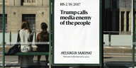 Ein Poster an einer Bushaltestelle zeigt den Slogan 'Trump calls media enemy of the people'. Daneben eine Glasscheibe, hinter der zwei Menschen sitzen mit dem Rücken zum Fotografen