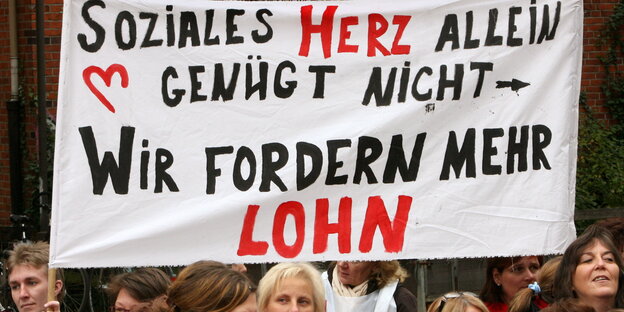 Mehrere Frauen demonstrieren, über ihren Köpfen hängt ein großes Tuch mit der Aufschrift "Soziales Herz allein genügt nicht - wir fordern mehr Lohn"