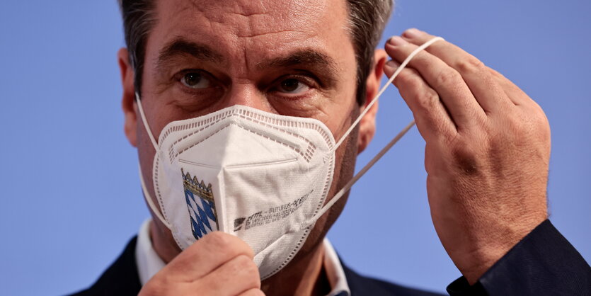 Markus Söder setzt sich eine FFP3-Maske auf. Die Maske hat ein Bayerische Wappen auf der Seite.