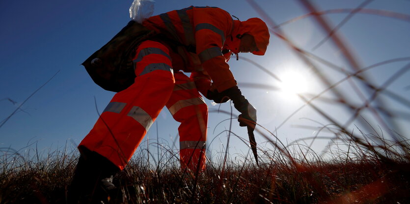 Ein Mann steht mit Leuchtanzug auf einem Moor mit einem Messer und einem Büschrel in der Hand