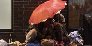 Zwei Personen sitzen am Boden und schützen sich mit einem roten Regenschirm