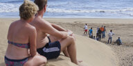 Ein Mann und eine Frau in Badekleidung beobachten am Strand von Gran Canaria ein Gruppe von Migranten