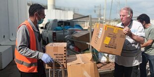 Zwei Männer entladen Pakete von einem Gepäckwagen, im Hintergrund die Mauer von Gaza