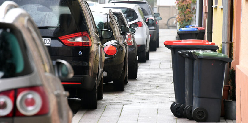 Autos parken auf einem Bürgersteig, der zusätzlich von Mülltonnen versperrt wird. Der Platz auf dem Gehweg ist eng