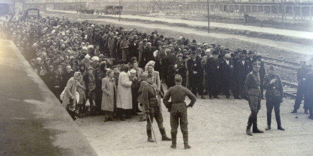 Das Konzentrationslager Auschwitz in Polen. Ein Bahnhof mit vielen Menschen, die - nach Geschlechtern getrennt - in Reihen stehen. Im Vordergrund NS-Personal.