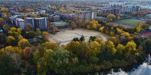 Ein sandiger Platz von Bäumen umringt, dahinter hohe Wohnhäuser.