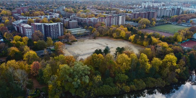 Ein sandiger Platz von Bäumen umringt, dahinter hohe Wohnhäuser.