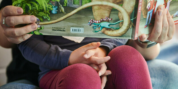 Hände einer Erzieherin halten ein Buch und Kind, das auf dem Schoss sitzt