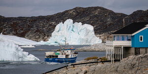 Ein Fischerboot fähr an einem Eisberg vorbei. Im Hintegrund eine Felsenküste.