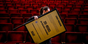 Das Bild zeigt eine Frau, die ein Schild mit dem Hinweis auf die 2G-Regel durch die Sitzreihe eines Theaters trägt.