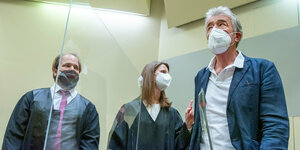 Drei Menschen mit Maske stehen im Gerichtssaal