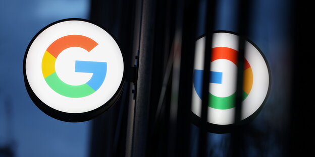 Es ist dunkel. An einer verglasten Hausfassade hängt ein leuchtendes "G" für "Google". Es spiegelt sich in der Fassade.