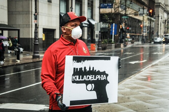 Ein Mann steht mit einem SChild auf einer Straße in Philadelphia, auf dem eine Pistole zusehen ist und die Aufschrift "Killadelphia"