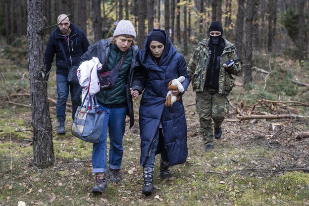 Eine junge Frau begleitet zwei Geflüchtete, Mann und Frau, durch den Wald. Im Hintergrund Grenzsoldatin in Tarnkleidung