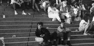 Schwarz-weiß-Fotografie. Zwei Punker sitzen auf einer Treppe. Umringt von Menschen.