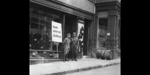 Zwei Männer der SA stehen vor einem Herrenbekleidungsgeschäft, hinter ihnen steht ein Schild: kauft nicht bei Juden