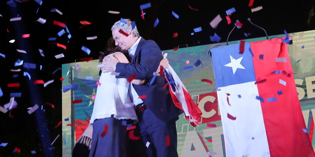Eine Frau und ein Mann umarmen sich, daneben die chilenische Flagge und viel Konfetti in den Farben der Flagge