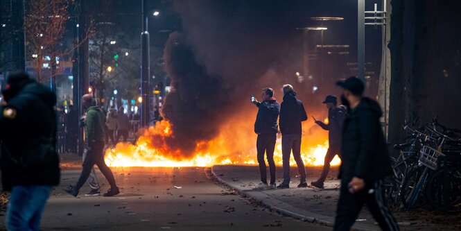 Eine Straße steht in Flammen, Menschen machen Handy-Aufnahmen oder gehen vorbei.