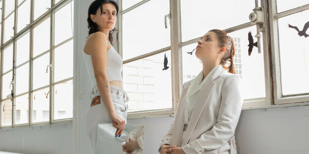 Zwei in Weiß gekleidete Performer:innen stehen und sitzen an einem Fenster. Die Frau links hält eine Spiegelplatte in der Hand