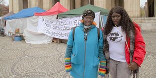 Zwei Frauen stehen vor einem langen beschrifteten Banner und Zelten