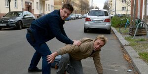 Ein Filmausschnitt zeigt zwei Männer auf einer Straße in Berlin. Einer kniet am Boden, der andere steht schützend über ihm.