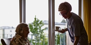 Eine Pflegerin bringt einer alten Frau ein Getränk und einen Kuchen.